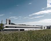 Modern white, low-profile building with a tall tower marked 112 set against a blue sky, edged by a cornfield in the foreground.