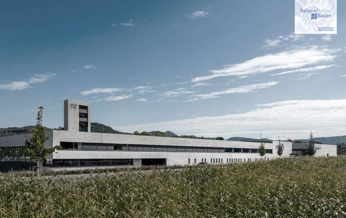 Modern white, low-profile building with a tall tower marked 112 set against a blue sky, edged by a cornfield in the foreground.