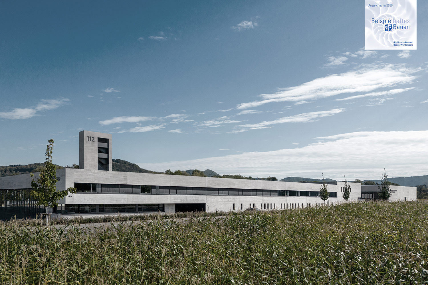 Neubau Feuerwehr und Bauhof Metzingen ausgezeichnet, Beispielhaftes Bauen-2026 | dasch zürn + partner Modern white, low-profile building with a tall tower marked 112 set against a blue sky, edged by a cornfield in the foreground.