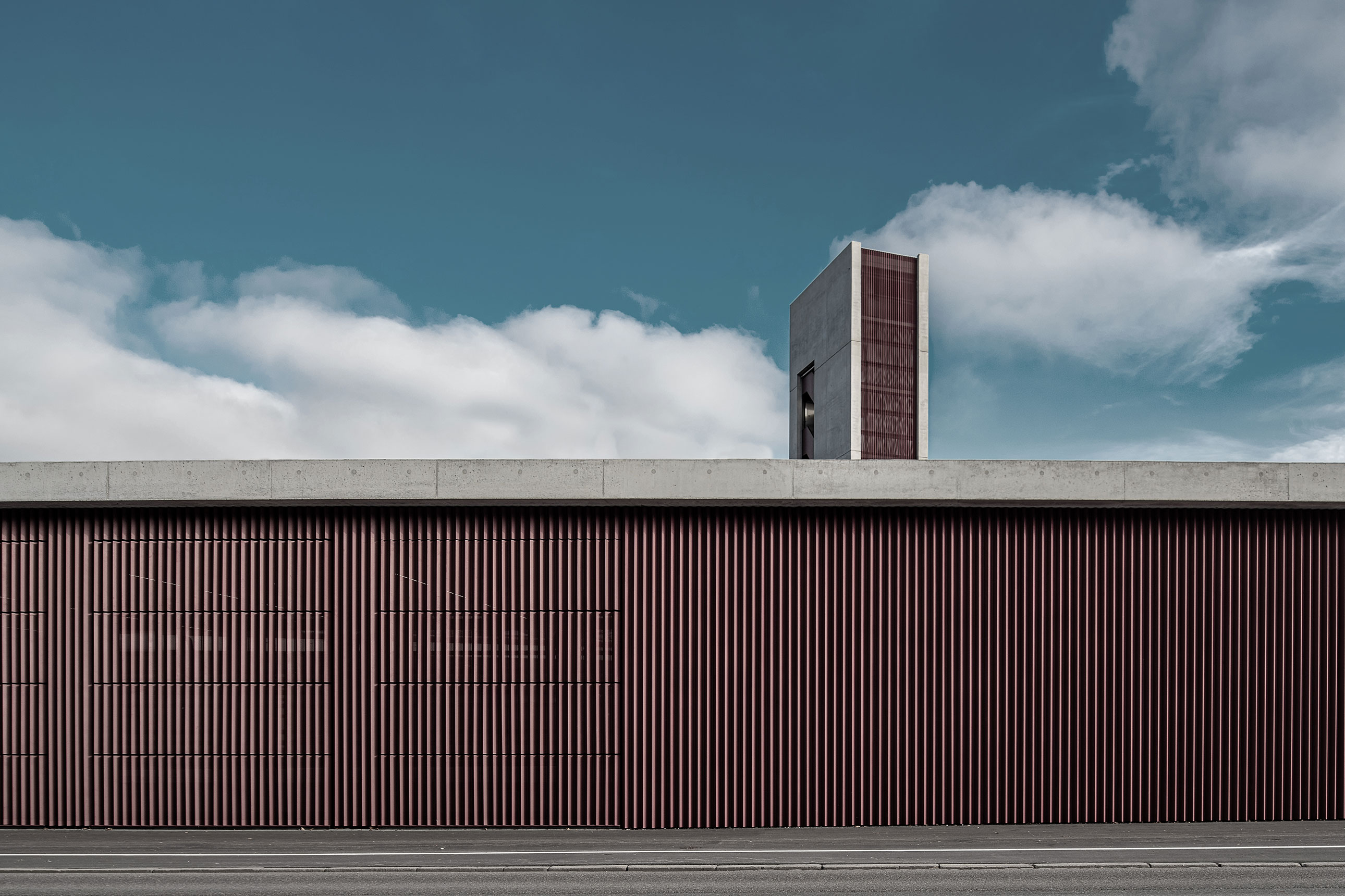 Modern concrete building with a tall red vertical louvered tower against a blue sky with clouds, foreground shows a flat road edge.